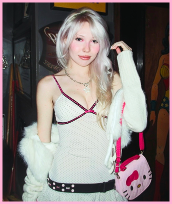 Young woman with long blonde hair (me) wearing a white top with black trim, a white fur coat, and a pink Hello Kitty bag. She stands in a dimly lit room.